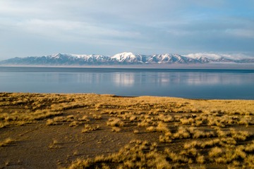 Snow-capped mountain range across a broad lake, with dry steppe grass in the foreground under a pale sky.